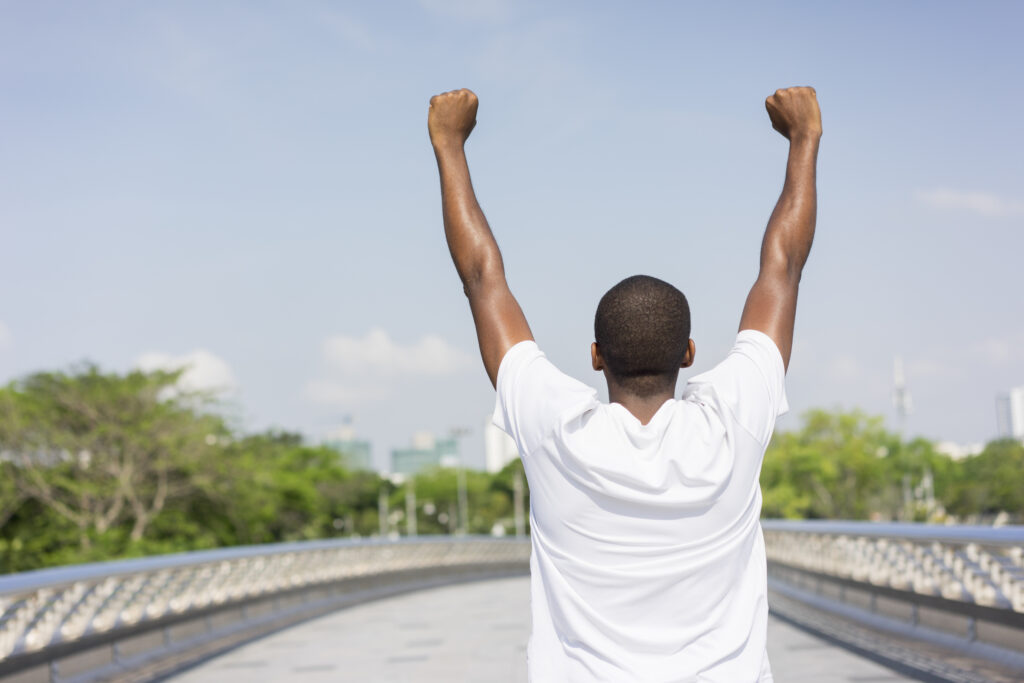 Back of black man enjoying city view and raising both hands. Young Afro American inspired by morning exercising outdoors. City sport and street workout concept